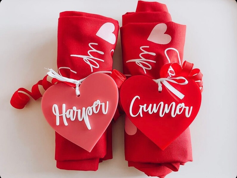 May include: Two rolled red napkins with white heart designs and the word "up" printed on them. Each napkin is adorned with a heart-shaped tag. One tag is a gradient pink with the name "Harper" in white, and the other is red with the name "Crumner" in white.