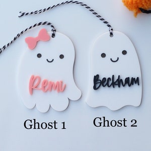 May include: Two white ghost-shaped tags with black and white string. One ghost has a pink bow and the name "Remi" in pink. The other ghost has the name "Beckham" in black. The text "Ghost 1" and "Ghost 2" are below the tags.