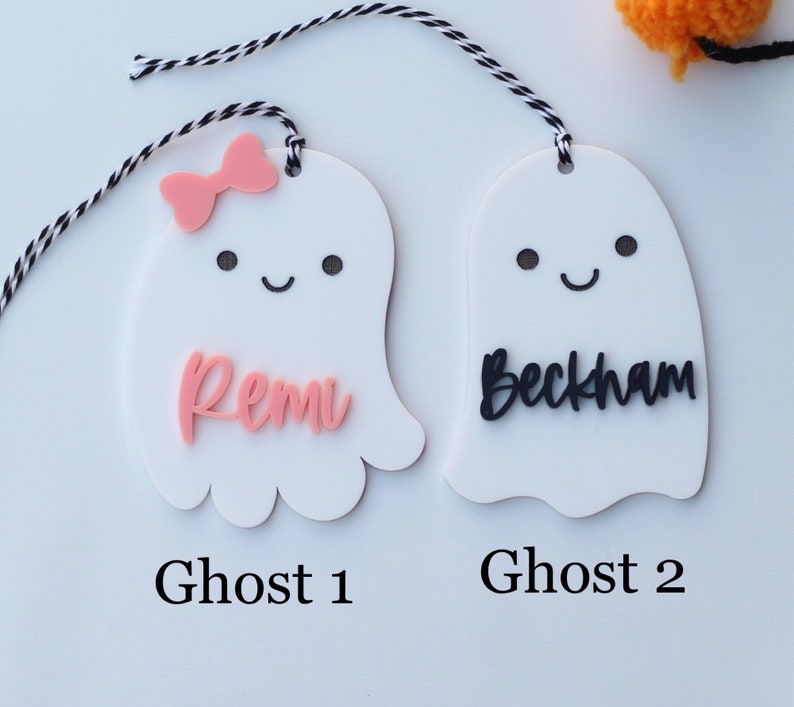 May include: Two white ghost-shaped tags with black and white string. One ghost has a pink bow and the name "Remi" in pink. The other ghost has the name "Beckham" in black. The text "Ghost 1" and "Ghost 2" are below the tags.