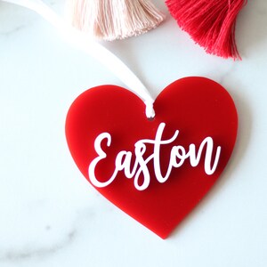 May include: A red heart-shaped ornament with the name "Easton" in white script. The ornament has a white ribbon for hanging. Decorative tassels in pink and red are visible in the background.