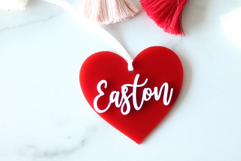 May include: A red heart-shaped ornament with the name "Easton" in white script. The ornament has a white ribbon for hanging. Decorative tassels in pink and red are visible in the background.