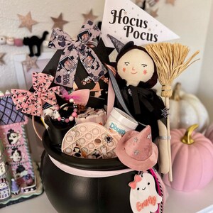 May include: A black cauldron filled with Halloween-themed items, including a witch doll, a broom, and a pink witch hat. The cauldron is decorated with bows and a ghost-shaped tag. A sign reads "Hocus Pocus".