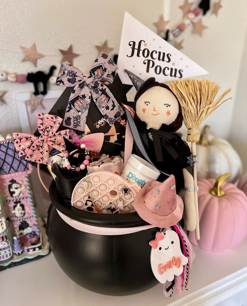 May include: A black cauldron filled with Halloween-themed items, including a witch doll, a broom, and a pink witch hat. The cauldron is decorated with bows and a ghost-shaped tag. A sign reads "Hocus Pocus".