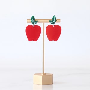 May include: A pair of red apple-shaped earrings with green leaves hanging from a gold earring stand.