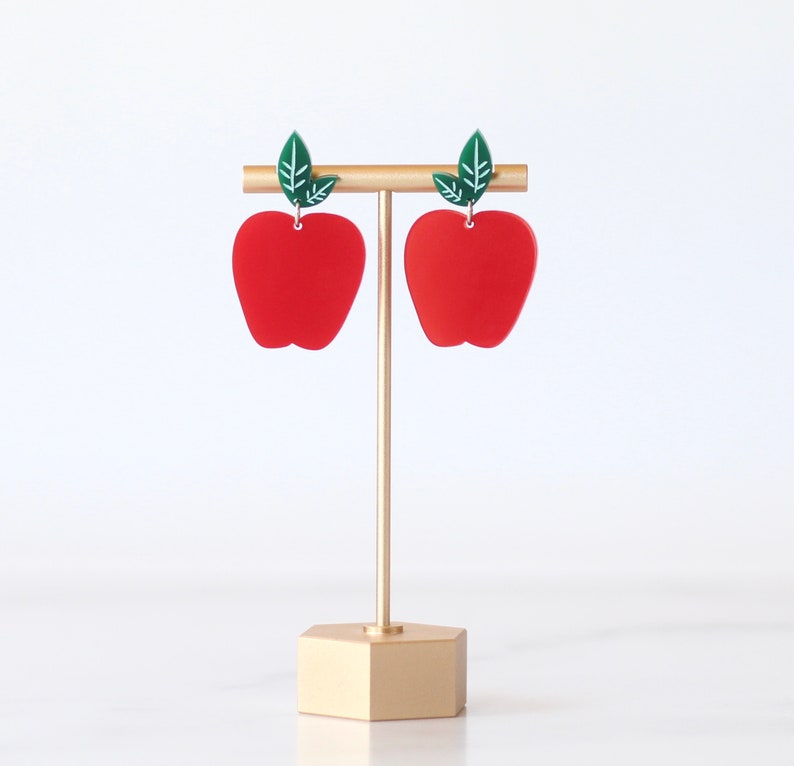 May include: A pair of red apple-shaped earrings with green leaves hanging from a gold earring stand.