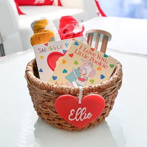 May include: A woven basket filled with baby items, including two children's books, a rattle, and a pair of red baby socks. A red heart-shaped tag with the name "Ellie" hangs from the basket.