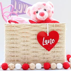 May include: A woven basket filled with a pink teddy bear, a pink and white patterned item, and a pink heart-shaped decoration. A red heart-shaped tag with the name "Lane" hangs from the basket. A garland of red, white, and beige pom-poms decorates the base.