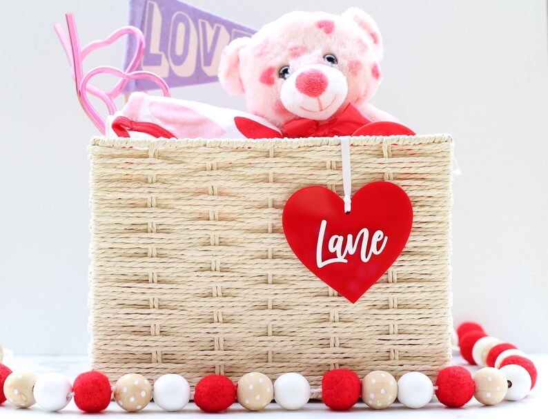 May include: A woven basket filled with a pink teddy bear, a pink and white patterned item, and a pink heart-shaped decoration. A red heart-shaped tag with the name "Lane" hangs from the basket. A garland of red, white, and beige pom-poms decorates the base.