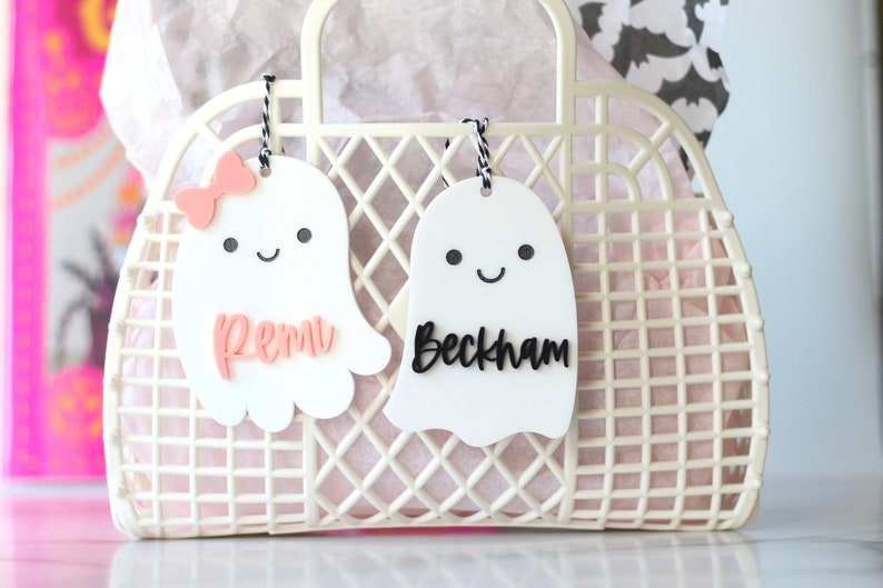 May include: A white plastic tote bag with a grid pattern, holding pink tissue paper. Two white ghost-shaped tags hang from the handle, one with a pink bow and the name "Remi", the other with the name "Beckham" in black.