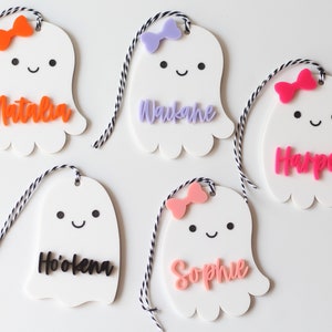 May include: White ghost-shaped name tags with colorful bows and names. Each tag has a smiling face and is strung with black and white twine. Names include Natalia, Waukane, Harper, Ho'okena, and Sophie. Perfect for Halloween or party favors.