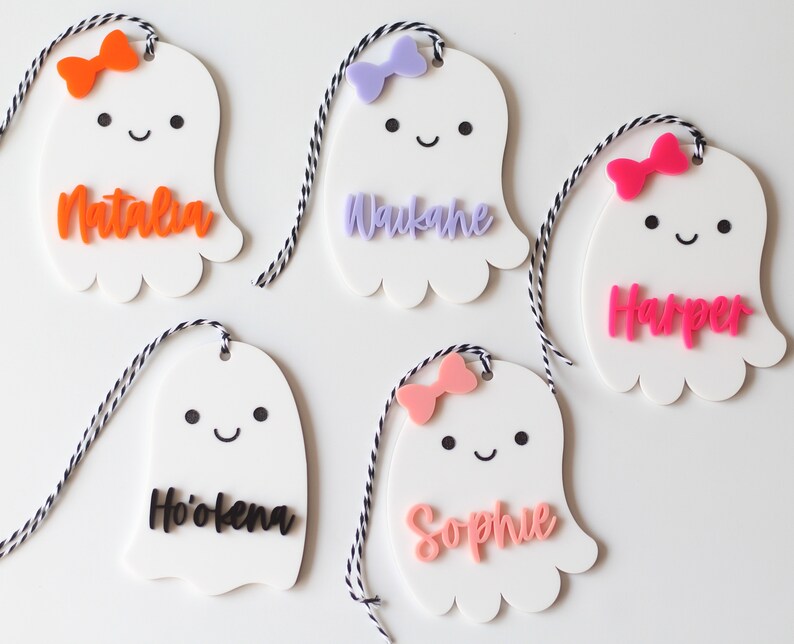 May include: White ghost-shaped name tags with colorful bows and names. Each tag has a smiling face and is strung with black and white twine. Names include Natalia, Waukane, Harper, Ho'okena, and Sophie. Perfect for Halloween or party favors.