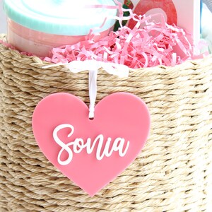 May include: A woven basket filled with pink shredded paper, a tumbler, and a book. A pink heart-shaped tag with the name "Sonia" in white script hangs from the basket. The tumbler is light blue and pink.