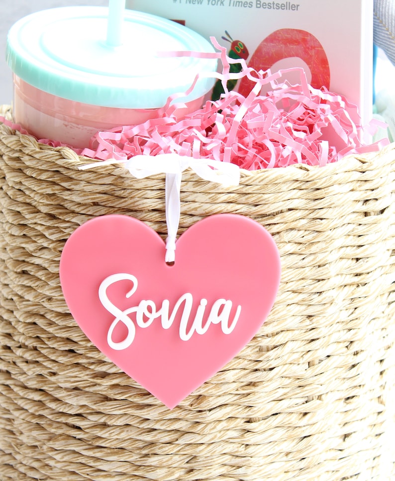 May include: A woven basket filled with pink shredded paper, a tumbler, and a book. A pink heart-shaped tag with the name "Sonia" in white script hangs from the basket. The tumbler is light blue and pink.