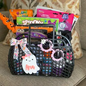 May include: A black plastic basket filled with Halloween-themed items. The basket contains orange and green glow sticks, nail polish, and decorations. A ghost-shaped tag with the name "Ava" hangs from the handle, along with a ribbon that says "No Tricks, Just Treats!" Pink sunglasses with the word "HAPPY" are attached.
