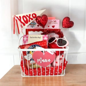 May include: A white wire basket filled with Valentine's Day gifts. Items include a pennant flag with "XOXO", heart-shaped sunglasses, heart-shaped decorations, and a tag that says "Maeve". The basket is on a wooden surface.