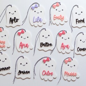 May include: White ghost-shaped name tags with black and pink names, each with a small bow and a black and white string. Names include Arlo, Lila, Emily, Ford, Mary Claire, Ana, Dalton, Ava, Cameron, Connor, Graham, Chloe, and Maddie.