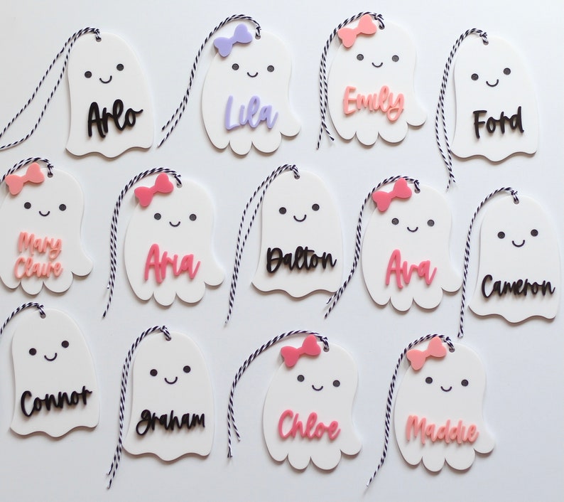 May include: White ghost-shaped name tags with black and pink names, each with a small bow and a black and white string. Names include Arlo, Lila, Emily, Ford, Mary Claire, Ana, Dalton, Ava, Cameron, Connor, Graham, Chloe, and Maddie.