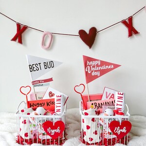 May include: Two white wire baskets filled with Valentine's Day gifts, including heart-shaped decorations and flags that read "Best Bud" and "Happy Valentines Day." A banner with "XOX" hangs above. Each basket has a heart-shaped name tag.