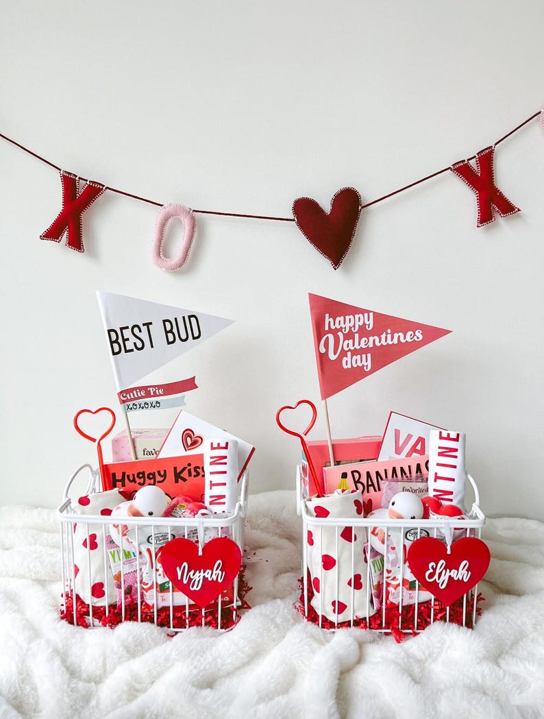 May include: Two white wire baskets filled with Valentine's Day gifts, including heart-shaped decorations and flags that read "Best Bud" and "Happy Valentines Day." A banner with "XOX" hangs above. Each basket has a heart-shaped name tag.