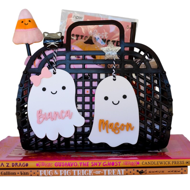 May include: A black plastic basket filled with Halloween-themed items, including a candy corn-shaped pen, a black cat charm, and two white ghost-shaped tags with names. The basket sits on top of a stack of children's books.