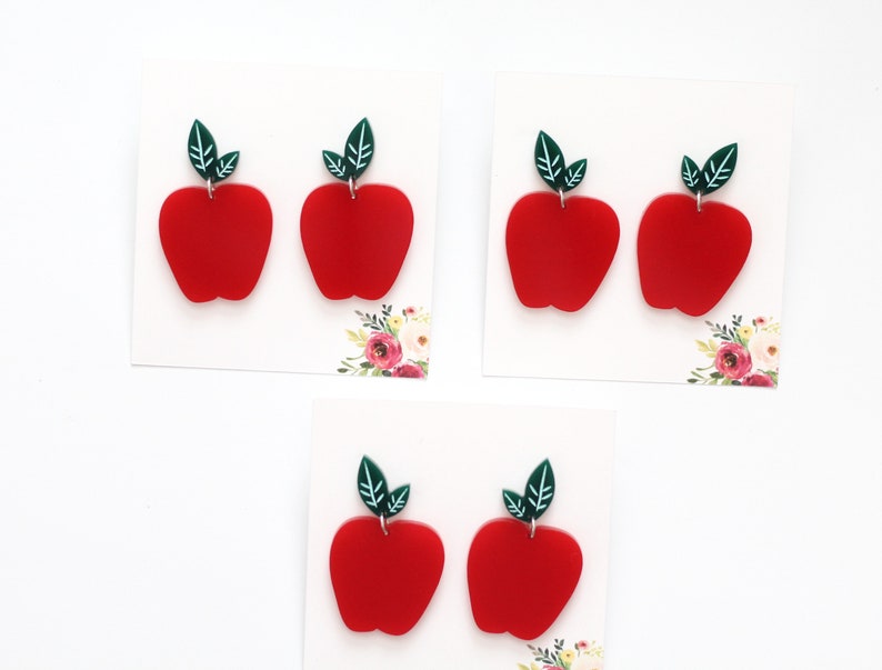 May include: A set of four red apple earrings with green leaves. The earrings are made of acrylic and are perfect for adding a touch of whimsy to any outfit.