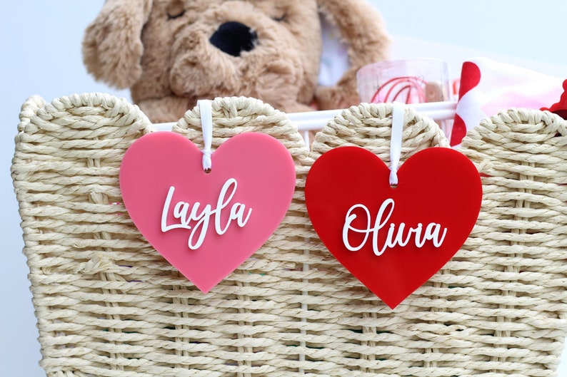 May include: Two heart-shaped gift tags, one pink and one red, with white script names "Layla" and "Olivia". The tags hang from a woven basket containing a stuffed animal and other gifts. The basket is made of light-colored woven material.