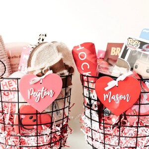May include: Two wire baskets filled with gifts, including a pink heart-shaped tag that says "Peyton" and a red heart-shaped tag that says "Mason". The baskets contain various items, such as a plush hat, a Funko Pop figure, and a red towel.
