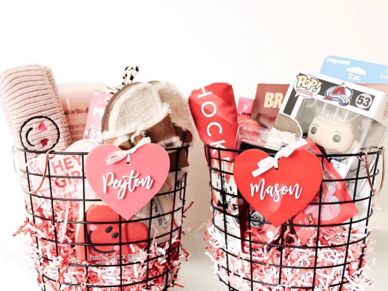 May include: Two wire baskets filled with gifts, including a pink heart-shaped tag that says "Peyton" and a red heart-shaped tag that says "Mason". The baskets contain various items, such as a plush hat, a Funko Pop figure, and a red towel.
