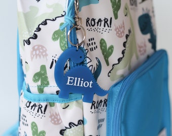 May include: Blue dinosaur-shaped keychain with the name "Elliot" printed in white. The keychain is attached to a blue backpack with a white dinosaur print.