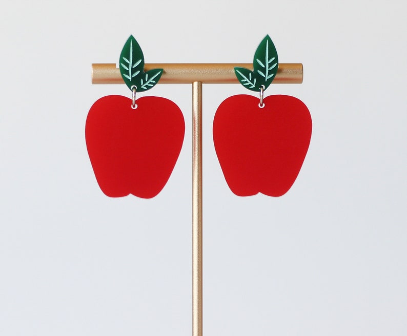 May include: A pair of red apple-shaped earrings with green leaf accents. The earrings are on a gold stand.