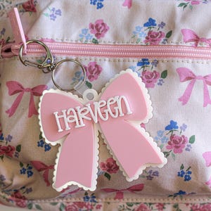 May include: A pink bow-shaped keychain with the name "HARVEEN" in white lettering. The keychain is attached to a floral patterned bag with pink bows, roses, and blue flowers. The bag has a pink zipper and a pink strap.