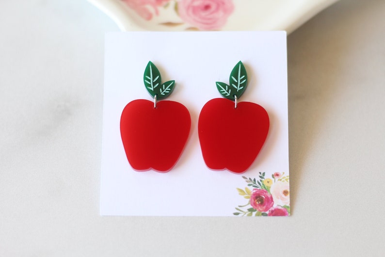 May include: A pair of red apple earrings with green leaves. The earrings are made of acrylic and have a glossy finish.