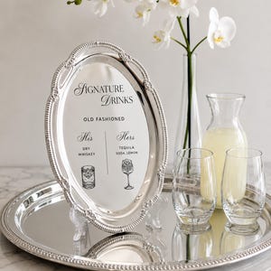 May include: An oval silver-toned tray with a beaded rim, displayed upright on a clear stand. The center features text reading Signature Drinks, Old Fashioned, His Dry Whiskey, and Hers Tequila Soda, Lemon, with illustrations of two different glasses.