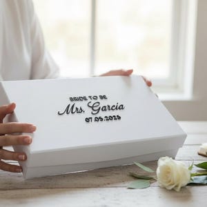 以下が含まれることがあります： 「BRIDE TO BE Mrs. Garcia 07.05.2025」の黒い文字が入った白いギフトボックス。白いバラとノートがテーブルにあります。箱は人が持っています。