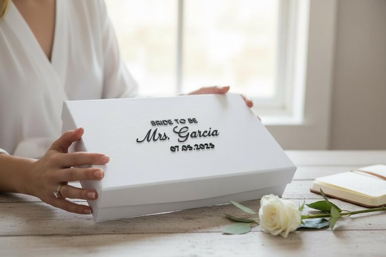 以下が含まれることがあります： 「BRIDE TO BE Mrs. Garcia 07.05.2025」の黒い文字が入った白いギフトボックス。白いバラとノートがテーブルにあります。箱は人が持っています。