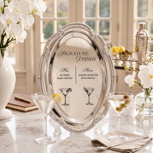 May include: An oval silver-toned serving tray with a decorative beaded rim, displaying the text SIGNATURE DRINKS, His LE NIMS Vesper Martini, and Hers MARIE-MARTINI Dirty Martini, accompanied by two martini glass illustrations and two filled glasses.