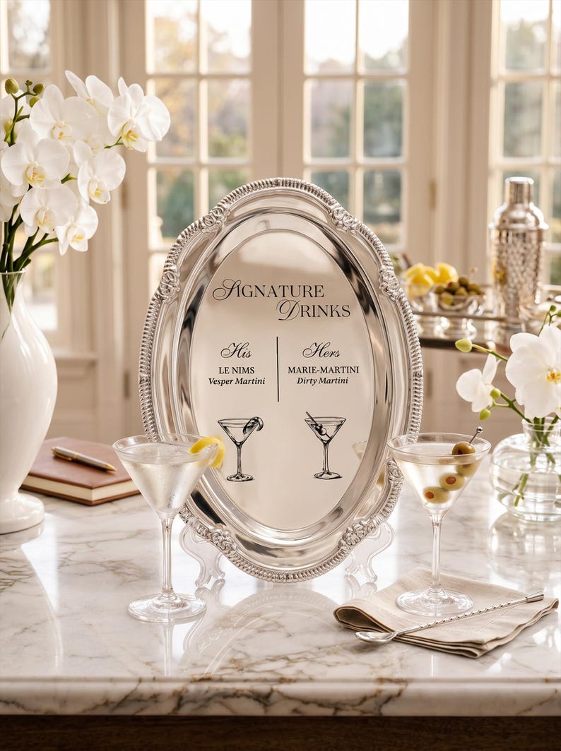 May include: An oval silver-toned serving tray with a decorative beaded rim, displaying the text SIGNATURE DRINKS, His LE NIMS Vesper Martini, and Hers MARIE-MARTINI Dirty Martini, accompanied by two martini glass illustrations and two filled glasses.