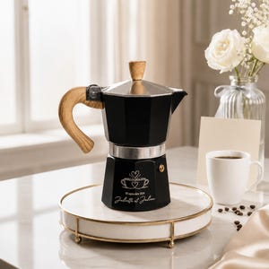 Op de afbeelding: Een zwarte moka-pot met een licht houten handvat en dekselknop. De basis is voorzien van een witte gravure van twee koffiekopjes met een hartje en de tekst 05 septembre 2026 en Juliette et Julien. De pot heeft een metallic zilveren middengedeelte.