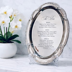 May include: An oval silver-toned metal tray with a decorative beaded edge, displayed on a clear stand. The center features a printed menu titled The Bar, listing various wines, beers, and spirits including Cabernet Sauvignon, Budweiser, and Tito's Vodka.