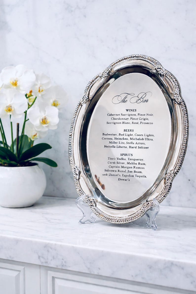 May include: An oval silver-toned metal tray with a decorative beaded edge, displayed on a clear stand. The center features a printed menu titled The Bar, listing various wines, beers, and spirits including Cabernet Sauvignon, Budweiser, and Tito's Vodka.