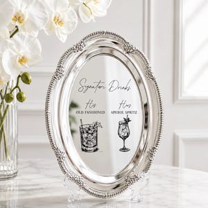 May include: An oval silver-toned mirror tray with a decorative beaded border. The center features black text reading Signature Drinks, His Old Fashioned, and Hers Aperol Spritz, accompanied by black line-art illustrations of an Old Fashioned and a cocktail glass.