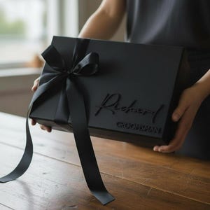 以下が含まれることがあります： 黒いギフトボックス。黒いリボンが結ばれ、蓋には「Richard」と「Groomsman」の文字がエンボス加工されています。ボックスは人が持っており、背景はぼやけています。