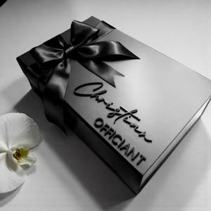 May include: A black gift box with a satin bow and the embossed text "Christian Officiant." A white orchid flower is in the lower left corner. The box is rectangular and has a shiny finish.