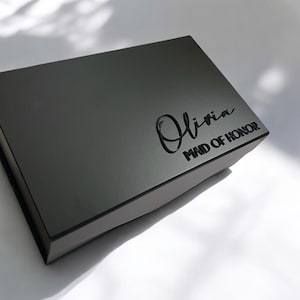 Può includere: Una scatola regalo grigio scuro con il nome "Olivia" in corsivo e le parole "MAID OF HONOR" in stampatello. La scatola ha una superficie liscia e riflettente.
