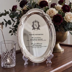 May include: An oval silver serving tray with an ornate embossed border, displaying a printed cocktail menu titled HOUSE FAVORITES. The text lists The Madame extra dirty martini and The Gentleman old fashioned with their respective ingredients.