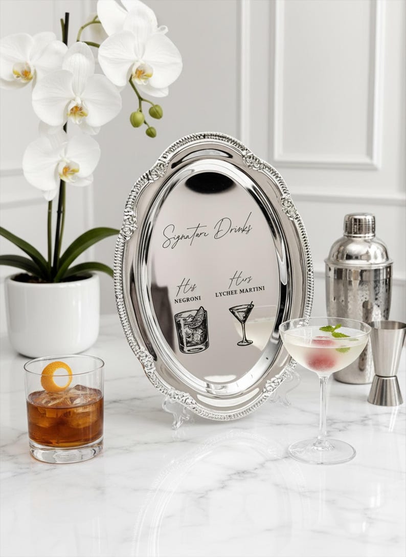 May include: An oval silver-toned metal tray with a decorative beaded rim, featuring the black printed text Signature Drinks, His Negroni, and Hers Lychee Martini, accompanied by illustrations of a cocktail glass and a rocks glass with ice.