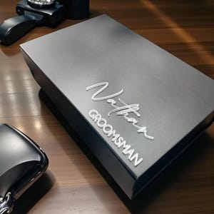 Może przedstawiać: Czarny pudełko na prezent z imieniem "Nathan" i słowem "Groomsman" w srebrnym skrypcie. Pudełko jest prostokątne i stoi na drewnianej powierzchni. Widoczne są inne czarne akcesoria.