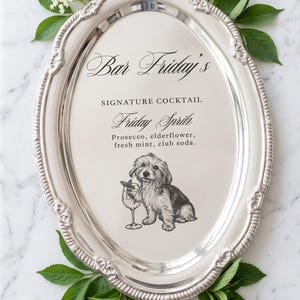 May include: An oval silver serving tray with an ornate rim featuring a printed menu for Bar Friday's Signature Cocktail, Friday Spritz, listing prosecco, elderflower, fresh mint, and club soda, accompanied by a black and white dog illustration.