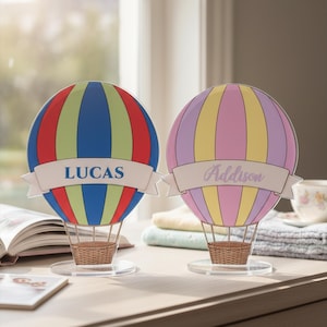 Peut inclure: Deux décorations en forme de montgolfière en matériau transparent. Le ballon de gauche présente des rayures verticales bleues, vertes et rouges avec une bannière blanche portant le nom LUCAS. Celui de droite a des rayures roses et jaunes avec Addison.