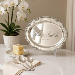 May include: An oval silver serving tray with a decorative beaded rim, engraved with the text "The Smiths" and "July 11, 2026". The tray is displayed on a clear stand, accompanied by a slice of cake on a plate and three silver spoons on a napkin.